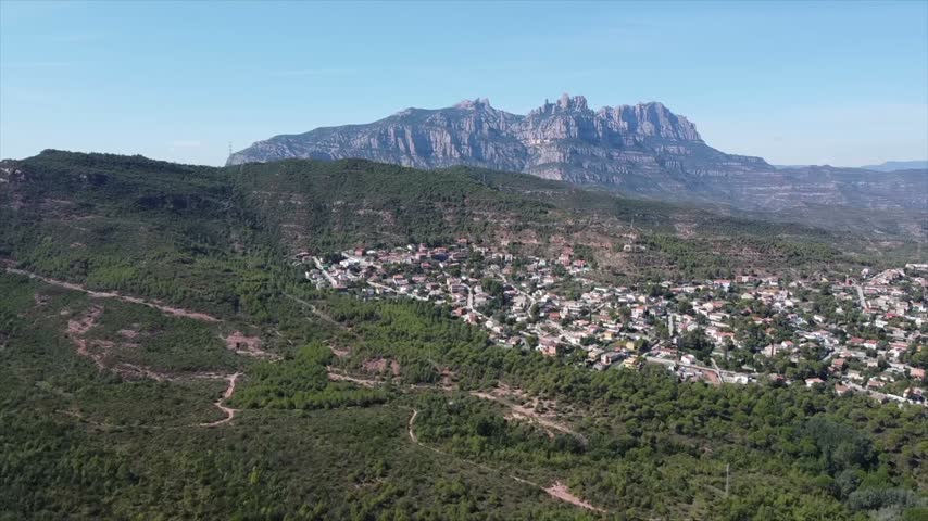 4K drone aerial view of Vacarisses, Catalonia, Spain. The green landscape surrounds the village, with the distinctive rocky peaks of Montserrat rising dramatically in the background.