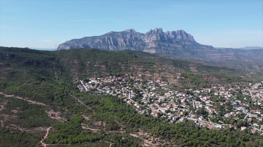4K drone aerial view of Vacarisses, Catalonia, Spain. The green landscape surrounds the village, with the distinctive rocky peaks of Montserrat rising dramatically in the background.