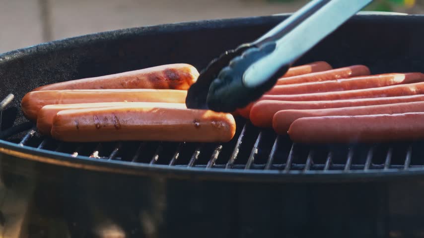 Tongs Flipping Hot Dogs Cooking on an Outdoor Barbecue Grill