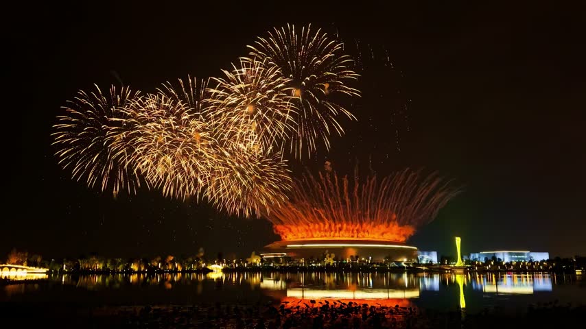 A vibrant fireworks display illuminates the night sky above a modern stadium and surrounding lake.