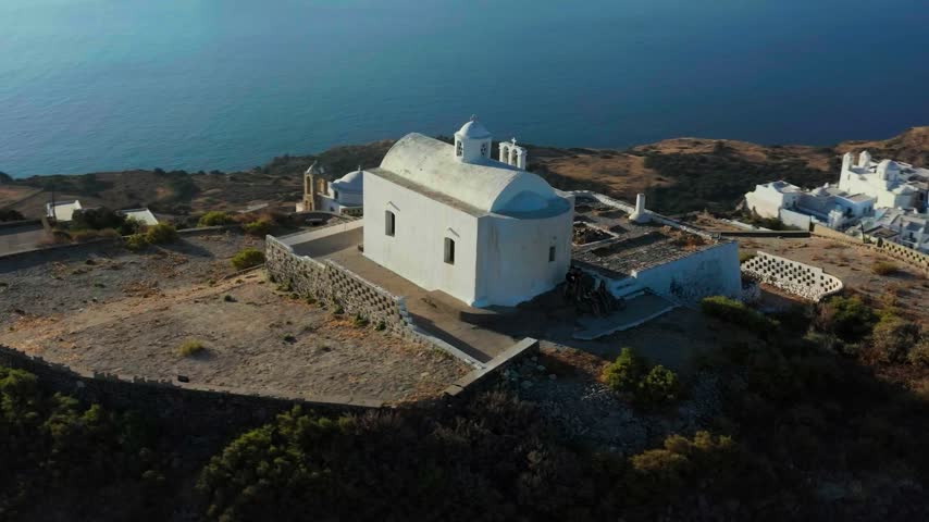 Cycladic village architecture and blue sea water surround a historic hilltop chapel.