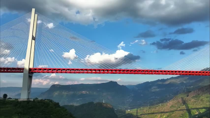 An impressive red cable-stayed bridge spans a deep, verdant mountain valley under a dramatic sky with white and dark clouds.
