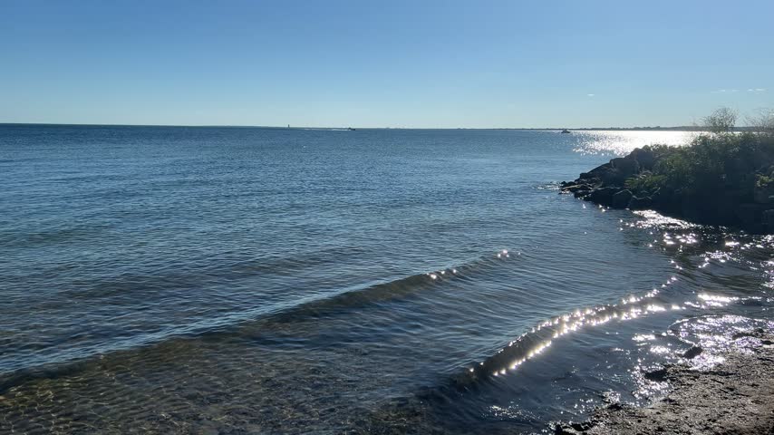 Gentle waves and sunlight sparkling on Lake St. Clair, Michigan, with a rocky shoreline, clear blue sky and boats on the horizon. Calm nature background.