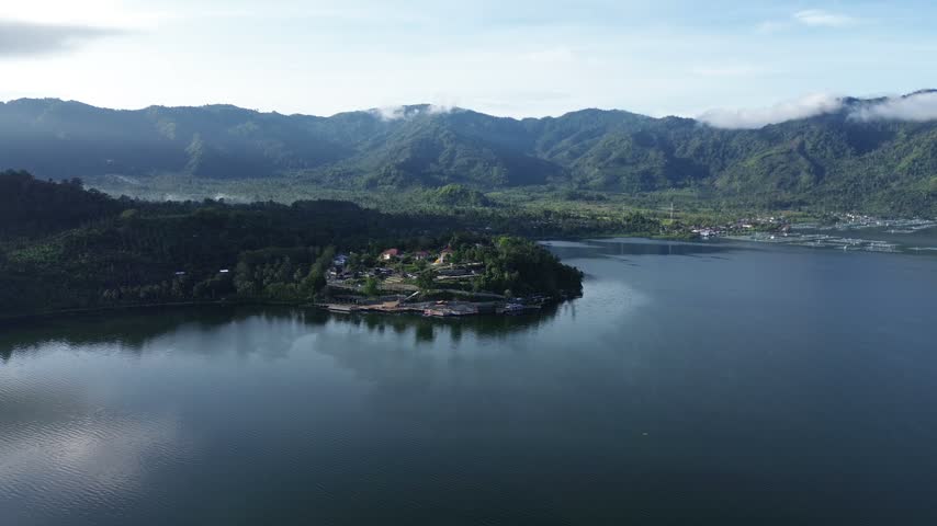 Panoramic aerial view of a scenic lake with a green peninsula, resort village, and majestic mountain ridges under daylight.