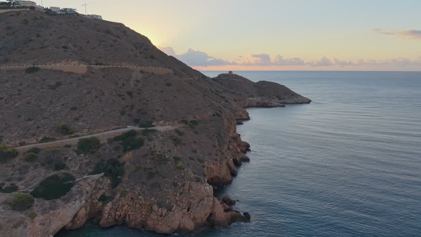 This clip showcases the coastline of Benidorm during sunset. The sun sets behind the cliffs, shining on the calm waters. Views highlight the natural beauty of the area.