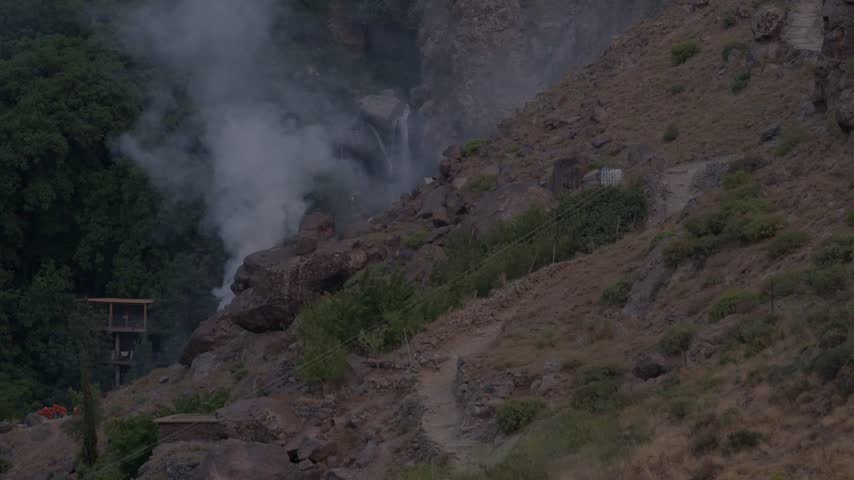 Cinematic shot of a rugged mountain gorge in the Moroccan Atlas range, showing thick smoke drifting past rocky hiking paths and lush ravine vegetation toward a distant hidden waterfall at dusk.