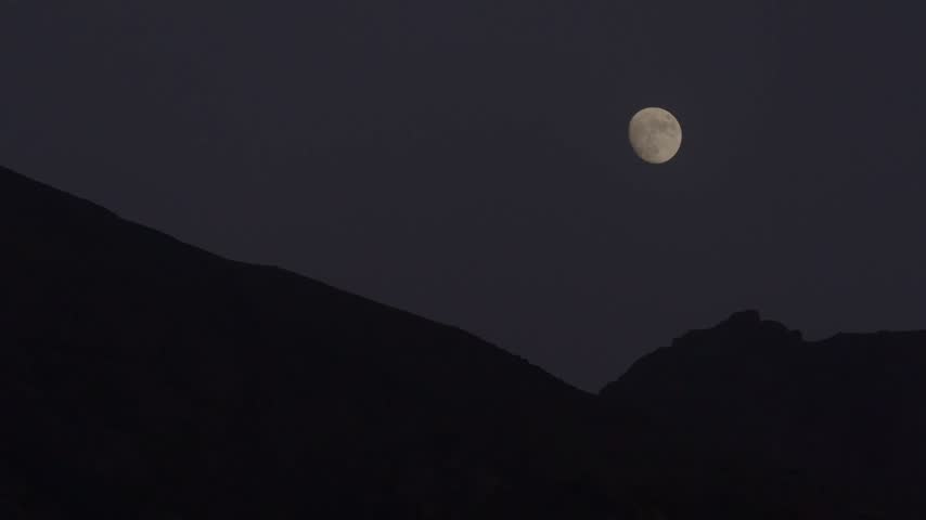 Cinematic night shot of a full moon rising over the dark, silhouetted mountain ridges of the High Atlas in Morocco, capturing a tranquil, mystical nocturnal landscape beneath a twilight sky.