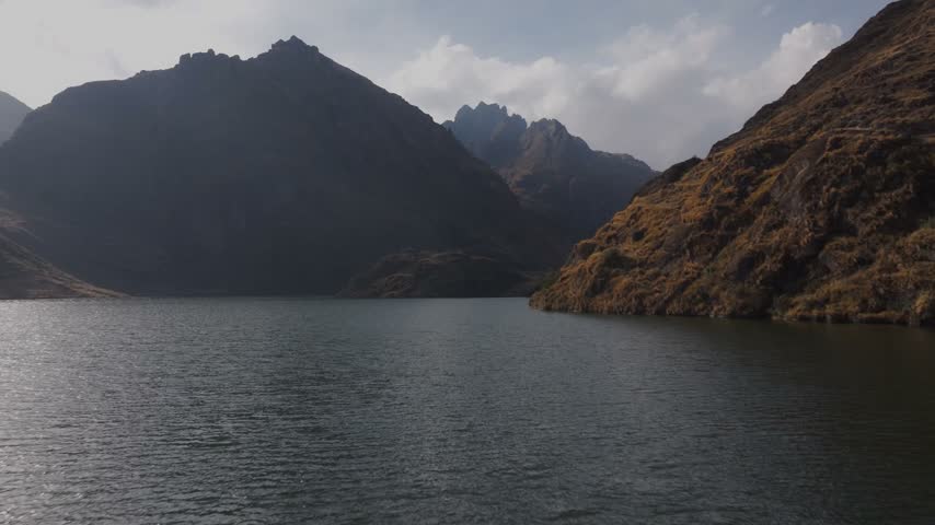 Aerial view skimming across a tranquil alpine glacial lake enclosed by towering, rugged mountain peaks and rocky slopes in the Peruvian Andes range, Peru.