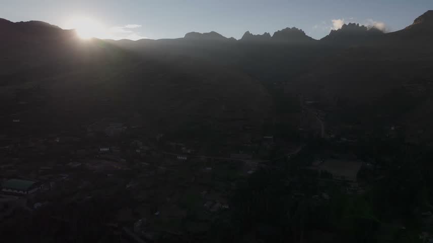 Aerial sunrise view over jagged Andean mountain peaks and a shaded high-altitude valley village in Peru, with bright sun rays breaking over the dark ridges.