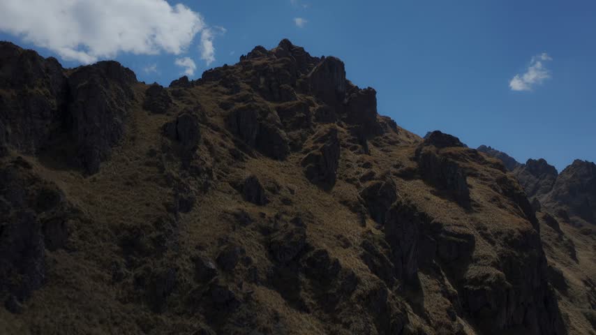 Cinematic aerial view of rugged rocky crags, jagged peaks, and dry Andean slopes rising beneath a deep blue sky with scattered clouds in the Andes of Peru.