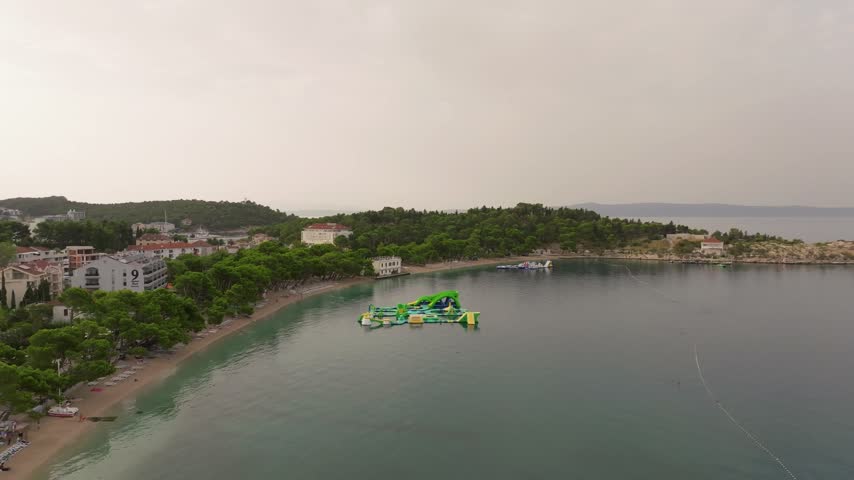 Inflatable aqua park floats off a quiet tree-lined beach near a small headland in Makarska under soft overcast skies.