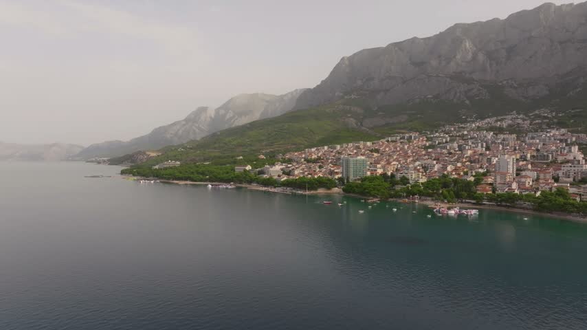 Seaside apartment blocks and waterfront park line a calm bay beneath a towering karst ridge in a quiet Croatian resort town.