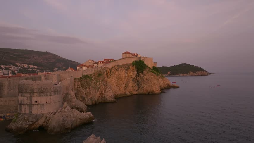 Coastal fortifications and red-roofed buildings sit on a rocky headland above calm water in soft twilight.