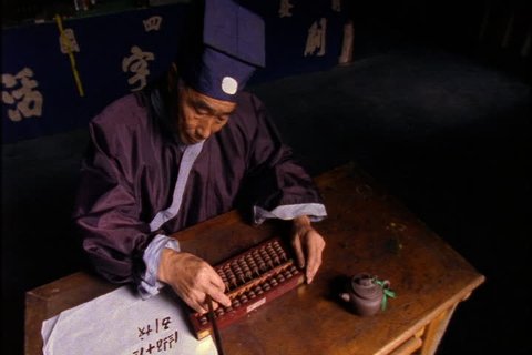 SONG DYNASTY VILLAGE, CHINA - NOVEMBER 09, 1999: Overhead view of man in traditional costume counting beads on an abacus and transcribing numbers onto a white scroll.