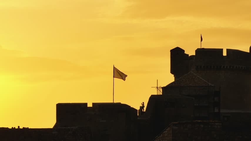 Dark castle ramparts and flags stand in silhouette against a rich orange evening sky as the sun drops below the horizon.
