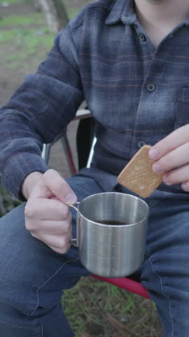 depicting a man wearing a red plaid flannel shirt and beanie, sitting in a snowy campfire setting and dipping a biscuit into a steaming blue enamel mug of hot coffee. Warm campfire glow, a steaming thermos, vintage lantern, and a tent surrounded by cold winter pine trees create a warm, adventurous atmosphere. Ideal for outdoor apparel branding, camping gear advertising