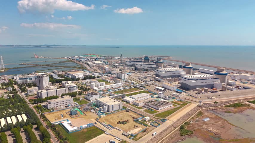 A large coastal industrial complex with various buildings and structures, adjacent to the sea under a clear sky.