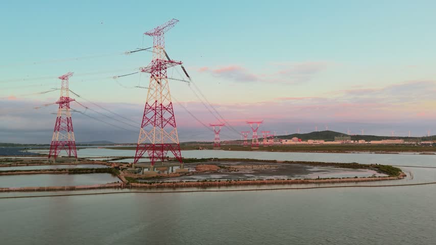 Red and white high-voltage transmission towers stand over water ponds at dusk, with distant hills in the background.