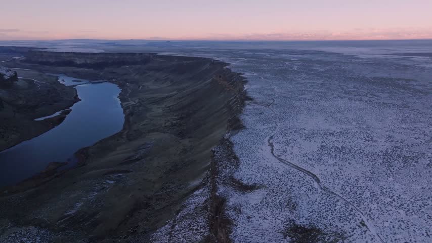 Canyon landscape featuring snow covered terrain along a steep rocky precipice adjacent to a winding body of water under a tranquil dawn atmosphere