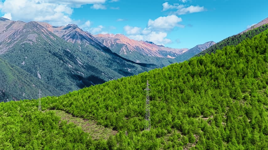 High voltage power transmission towers stand amidst lush green forest slopes with rugged mountain peaks under a blue sky.