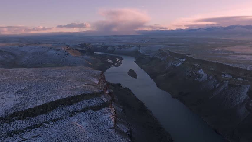 Canyon landscape featuring a winding river flowing past steep cliffs and snow dusted terrain under a vibrant morning sky during aerial drone capture