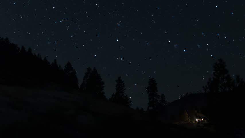 Star trails rotate across the clear dark sky above a silhouette of evergreen trees and hilly landscape in timelapse