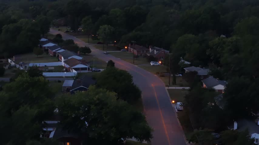 Two rows of small houses face each other across a Lynchburg street in Virginia at dusk, porch lamps lit, cars in the driveways and trees turning to silhouettes. Aerial lateral wide shot