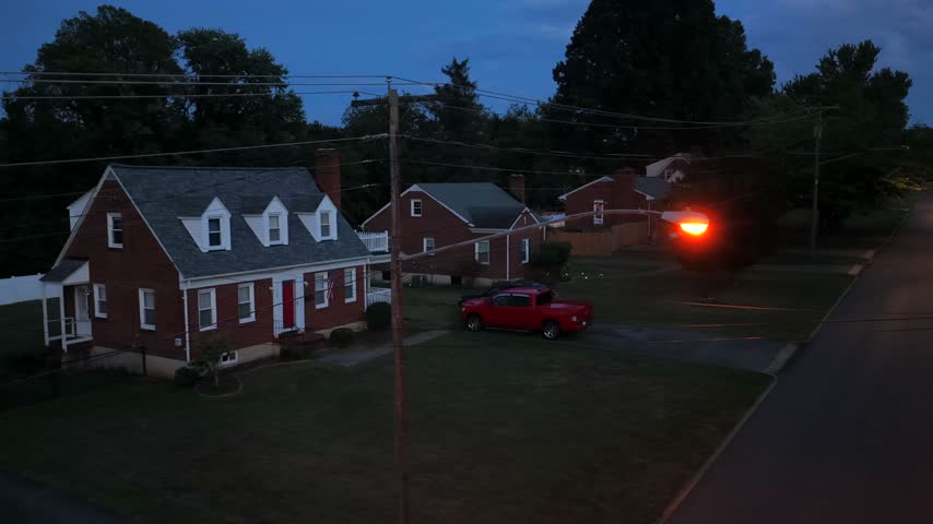 A red brick house with dormer windows and a red pickup in the driveway sits under a glowing street lamp in Lynchburg, Virginia, at blue hour. Aerial drone wide shot