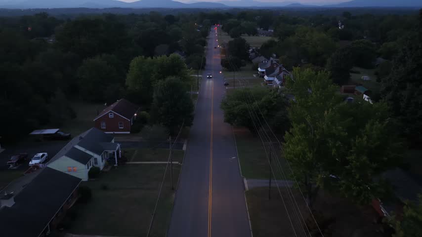 Modest brick and clapboard houses line a straight street in Lynchburg, Virginia, under a cloudy dusk sky, with power lines, mature trees and distant blue mountains. Aerial forward wide shot