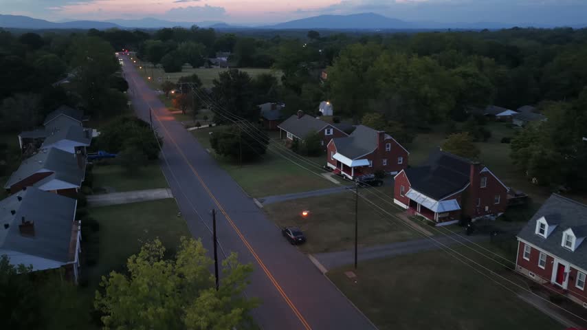 Red brick homes with white porches sit along a two lane road in Lynchburg, Virginia, at dusk, with lawns, pickup trucks and the Blue Ridge beyond the rooftops. Aerial lateral wide shot