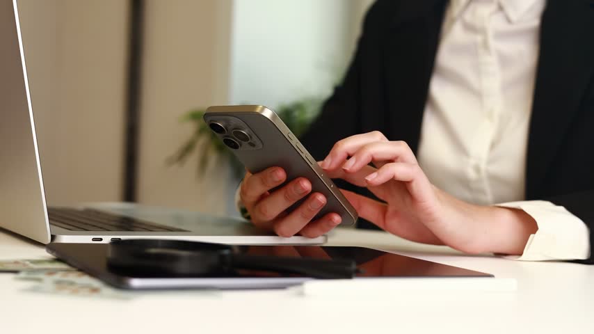 A person is using a phone while sitting at a desk with a laptop. Cash flow icons are visible on the screen. The setting is bright and suitable for work tasks.