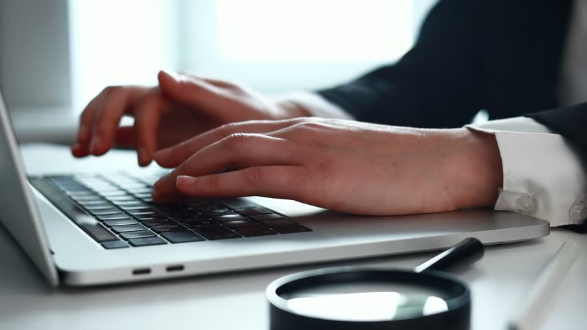 A person is typing on a laptop in an office. Bright light comes through a window. Around the screen, there are icons related to security and protection.