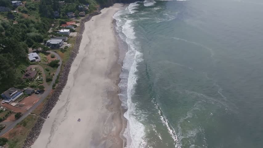 Oregon scenic coastline featuring sandy shores crashing waves forested cliffs and residential houses captured from an aerial perspective