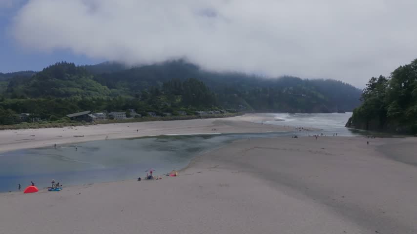 Oregon coastal landscape from above displaying a river emptying into the sea surrounded by sandy shores dense green forests and misty mountains under an overcast sky