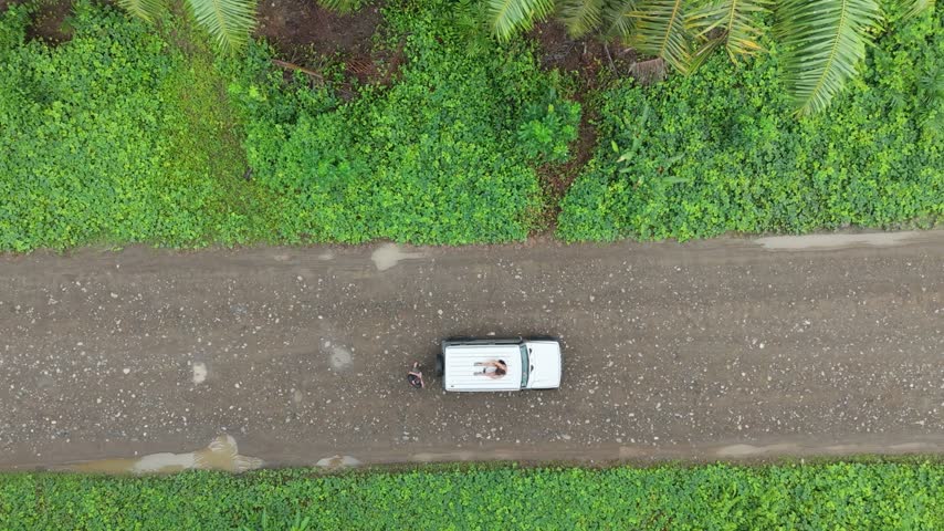 Aerial view of a white vehicle driving along a dirt road surrounded by lush green tropical vegetation and palm trees in Costa Rica.