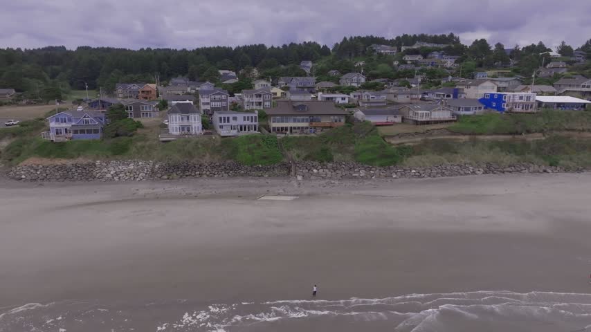 Oregon coastline featuring residential cliffside homes above a sandy beach with rolling waves and people strolling near the water under an overcast sky