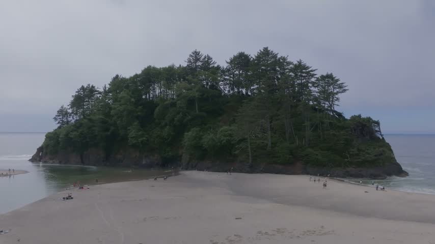Oregon coastline featuring verdant forested rocky islet joined to wide sandy shore by sandbar with distant ocean waves and vacationers enjoying leisure time