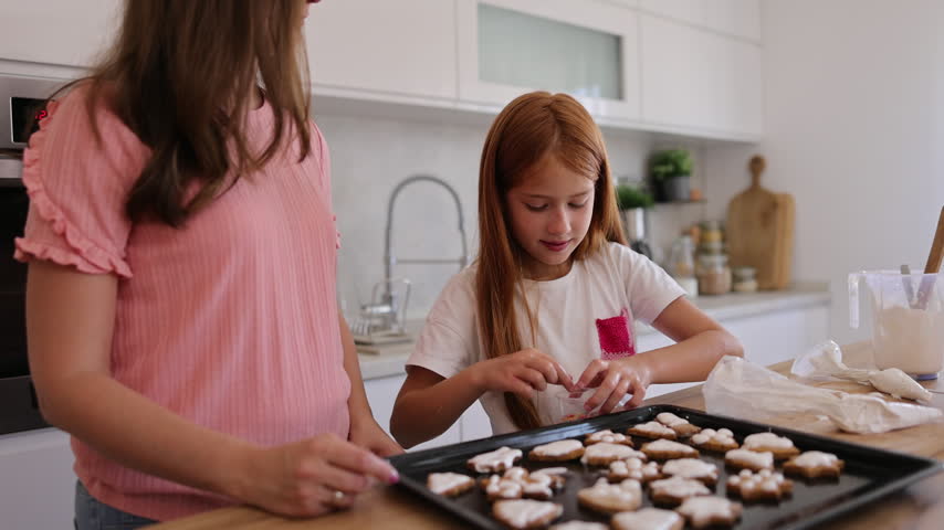 Mother and daughter having fun baking Christmas cookies together in the kitchen, adding colorful sprinkles over cookies coated in royal icing