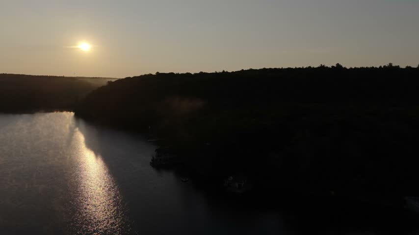 Mist drifts over a lake at sunrise near Muskoka, Ontario, drone advancing past tree shadows