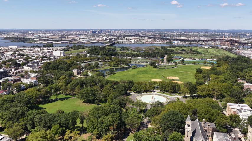 An aerial glide crosses a park in Jersey City, New Jersey, showing a circular fountain, baseball fields, tree-lined paths and surrounding neighborhoods, with waterways and bridges in the distance.