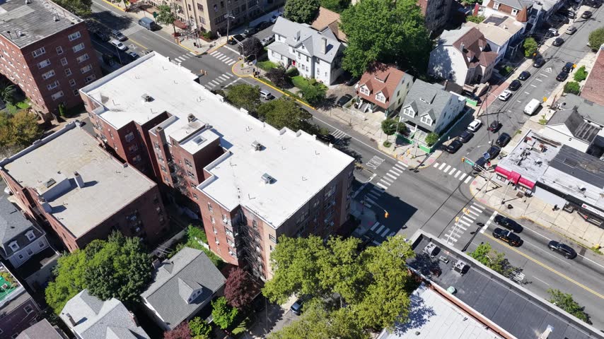 A drone pans across brick apartments and a street intersection in Jersey City, New Jersey, revealing white rooftops, leafy trees, crosswalks and closely spaced homes in a sunny urban neighborhood.