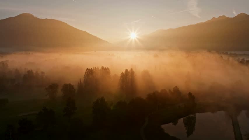 Morning sun beams shine through thick fog over a forest valley nestled between high mountains at dawn creating a peaceful and scenic atmosphere with warm natural light tones in this wide outdoor view