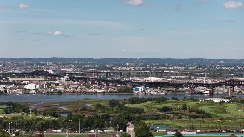 A drone pan follows river bridges beyond green parkland in Jersey City, New Jersey, showing transport corridors, distant industrial buildings and a scenic urban horizon above tree-covered grounds.