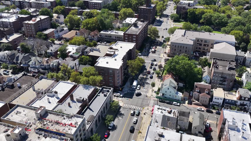 A drone pans over Jersey City, New Jersey, revealing tree-lined streets, brick apartment blocks, road intersections and tightly spaced homes as the view opens across a sunlit residential district.