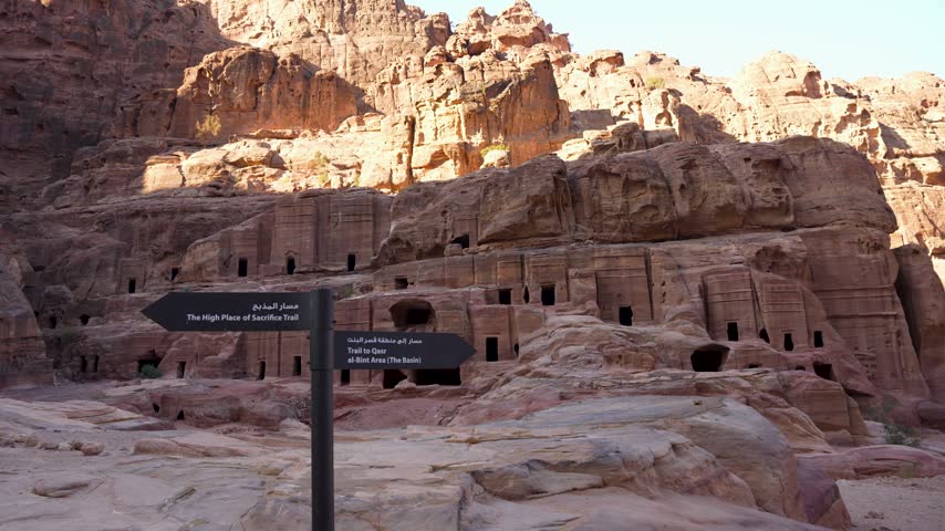 Sandstone rock-cut tombs and walking trails at Petra, Jordan, with directional signs leading visitors through the ancient Nabataean archaeological site, smooth handheld panning view