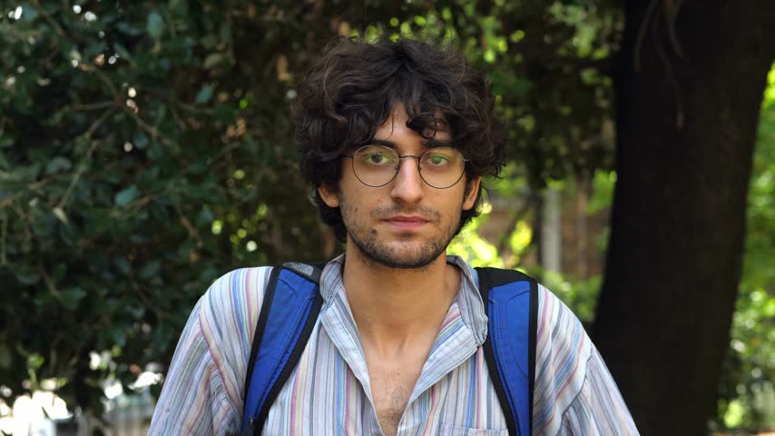 A young man with curly short hair and a light shirt, wearing glasses, looks seriously at the camera while standing in a park, capturing a moment of contemplation and focus.