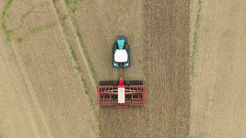 Tractor with seed drill working across cultivated farmland during crop planting season. Agricultural machinery preparing soil for efficient sowing. Modern agribusiness and mechanized farming on large