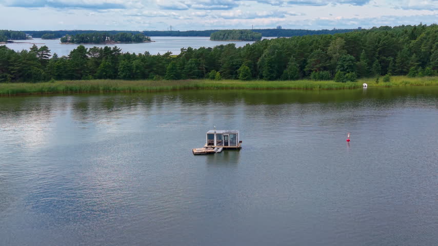 Floating sauna on a calm Baltic Sea bay in Finland, surrounded by pine forests and reed beds under a partly cloudy summer sky