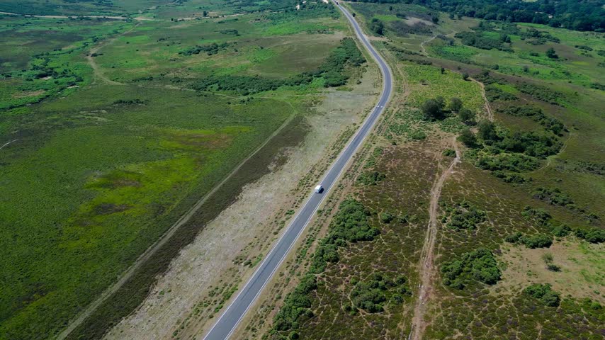 Aerial view showing single car traveling along winding road through open heathland with scattered greenery and rural terrain across New Forest Hampshire England in summer
