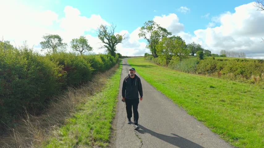 Countryside traveler with backpack walking along winding rural road under bright blue sky. Middle aged man hiking between green hedgerows and open fields. Outdoor fitness and active lifestyle during